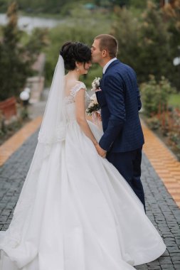 Tuğla duvarın önünde bir gelin ve damat öpüşüyor. Gelin beyaz bir elbise giyiyor ve damat mavi bir takım giyiyor.