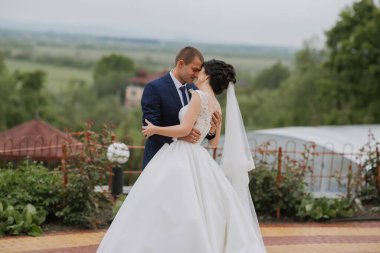 Bir gelin ve damat bir bahçe ortamında birbirlerini kucaklıyorlar. Gelin beyaz bir elbise giyiyor ve damat mavi bir takım elbise giyiyor. Sahne romantik ve samimi.