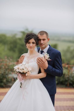 Gelin ve damat bir resim için poz veriyor, gelin bir buket çiçek tutuyor. Sahne mutlu ve romantik, çünkü çift düğün günlerini kutluyor.