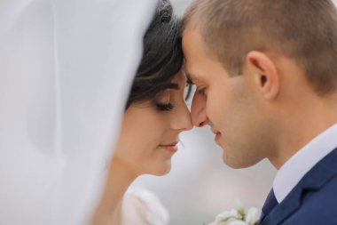 Bir gelin ve damat, yüzleri birbirine yakın bir şekilde birbirlerini kucaklıyor. Gelin beyaz bir peçe takıyor ve damat mavi bir takım elbise giyiyor. Çiftler arasındaki sevgi ve yakınlık kavramı