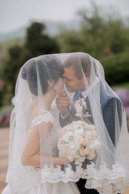 Bir gelin ve damat peçenin altında öpüşüyor. Gelin beyaz bir elbise giyiyor ve damat mavi bir takım elbise giyiyor. Gelin elinde bir buket çiçek tutuyor.