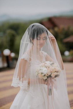 Bir gelin peçe takıyor ve elinde bir buket çiçek tutuyor. Duvak beyaz ve yüzünü örtüyor. Ona gizemli ve zarif bir görünüm veriyor.