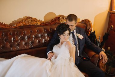 Gelin ve damat bir kanepede oturuyor, gelin damada yaslanıyor. Kanepe kahverengi ve çift resmi olarak giyinmiş. Sahne romantik ve samimi.