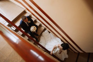 Gelin ve damat, beyaz bir elbise giymiş gelinle merdivenlerde dikiliyorlar. Adam elinde bir buket çiçek tutuyor ve kadın da elinde bir buket çiçek tutuyor.