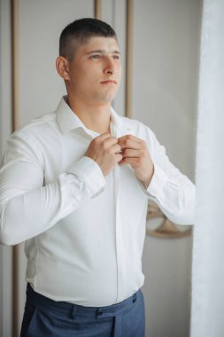 Beyaz gömlekli bir adam giyiniyor. Mavi pantolon giyiyor ve sakalı var.