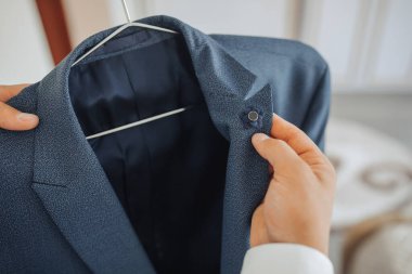 Bir adam elinde gümüş askılı mavi bir ceket tutuyor. Ceket parlak bir maddeden yapılmış ve üzerinde bir desen var. Adam cekete faiziyle bakıyor.