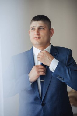 Mavi takım elbiseli bir adam kravatını bağlamaya hazırlanıyor. O, elindeki işe odaklanmış ve ciddi bir ruh hali içinde.