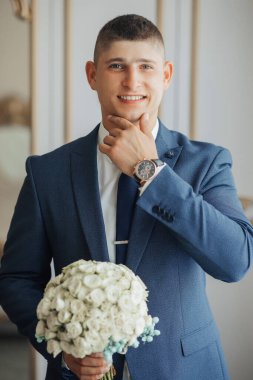 Takım elbiseli ve kravatlı bir adam elinde bir buket çiçek tutuyor. Gülümsüyor ve mutlu.