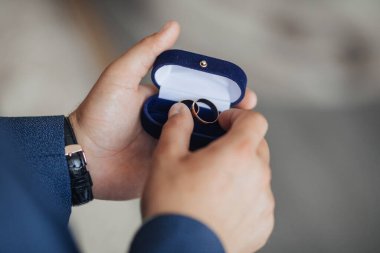 Bir adam içinde iki yüzük olan mavi bir kutu tutuyor. Adam takım elbise ve kravat takıyor.