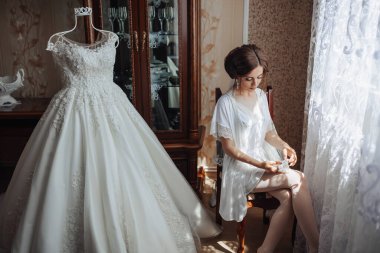 Bir kadın düğün için hazırlanıyor ve bir şifoniyerin önünde oturuyor. Arkasındaki duvarda uzun beyaz bir elbise asılı.