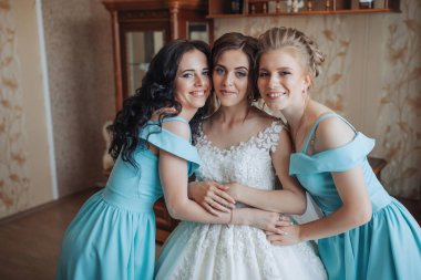 Üç kadın fotoğraf için poz veriyor, biri gelinlik giyiyor. Sahne mutlu ve kutlama dolu. Kadınlar gülümsüyor ve birbirlerine sarılıyorlar.