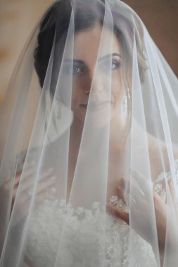 Bir gelin, kısmen yüzünü örten bir peçe takıyor. Duvak beyaz ve dantel bir tasarımı var. Gelin gülümsüyor ve mutlu.