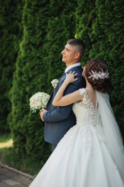 Bir gelin ve damat yeşil bir çitin önünde duruyor. Gelin beyaz bir elbise giyiyor ve elinde bir buket tutuyor. Damat mavi bir takım elbise ve kravat takıyor. İkisi de gülümsüyor.