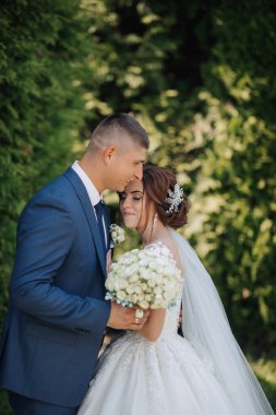 Bir gelin ve damat ormanda birbirlerine sarılıyorlar. Gelin elinde bir buket beyaz çiçek tutuyor.