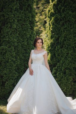 Beyaz elbiseli bir kadın yeşil bir çalılığın önünde duruyor. Kolye ve küpe takıyor.