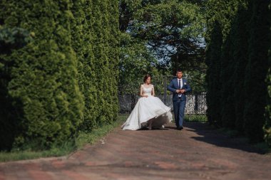 Bir gelin ve damat parkta bir patikada yürüyorlar. Gelin beyaz bir elbise giyiyor ve damat mavi bir takım elbise giyiyor. Çift mutlu görünüyor ve birlikte vakit geçiriyorlar.