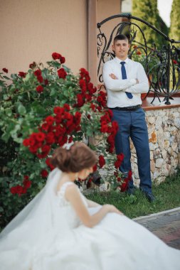 Bir gelin ve damat kırmızı güllerden oluşan bir çalılığın önünde duruyorlar. Gelin yerde oturuyor, ve damat da ona bakıyor. Sahne romantik ve samimi.