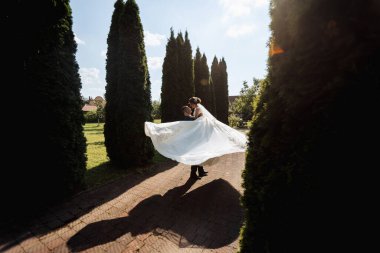 Gelin ve damat arka planda ağaçlar olan bir bahçede kucaklaşıyorlar. Gelin beyaz bir elbise tutuyor ve damat takım elbise giyiyor. Sahne romantik ve mutlu.