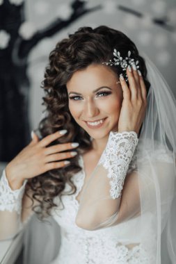 Uzun saçlı ve duvağı olan bir kadın gülümsüyor. Dantel bir elbise ve kolye giyiyor.