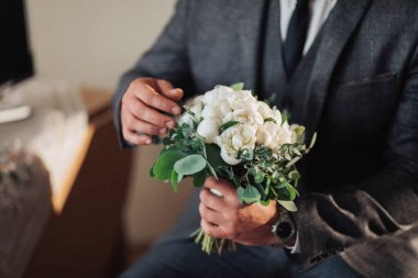 Takım elbiseli bir adam elinde bir buket beyaz çiçek tutuyor. Adam kravat takıyor ve kanepede oturuyor.