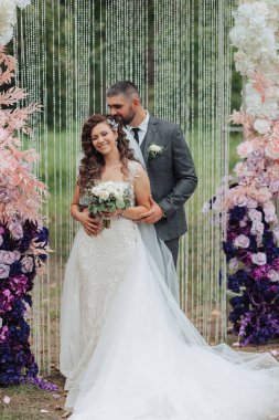 Bir gelin ve damat pembe bir kemerin önünde poz veriyorlar. Gelin bir buket tutuyor ve damat kravat takıyor. Sahne mutlu ve kutlama dolu, çünkü çift evleniyor.