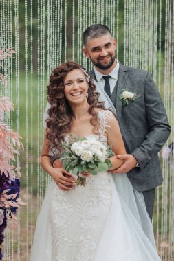 Bir gelin ve damat perdenin önünde poz veriyorlar. Gelin bir buket tutuyor ve damat kravat takıyor. Sahne mutlu ve kutlama dolu, çünkü çift evleniyor.