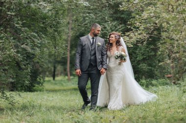 Bir gelin ve damat el ele tutuşarak ormanda yürüyorlar. Gelin beyaz bir elbise giyiyor ve damat da takım elbise giyiyor.