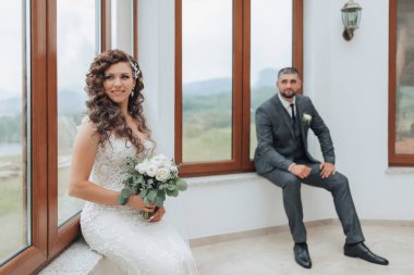 Bir gelin ve damat pencerenin önünde fotoğraf için poz veriyorlar. Gelin bir buket tutuyor ve damat bir takım elbise giyiyor. Sahne mutlu ve romantik.