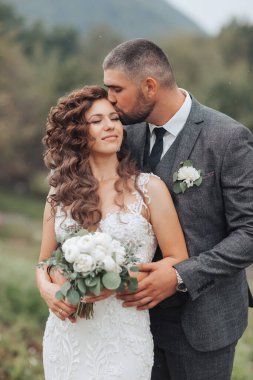 Bir gelin ve damat öpüşüyorlar ve gelin bir buket çiçek tutuyor. Sahne romantik ve mutlu.