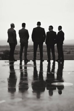 Takım elbiseli bir grup adam köprüde durup bir tarlaya bakıyor. Görüntünün kasvetli ve yansıtıcı bir havası var.