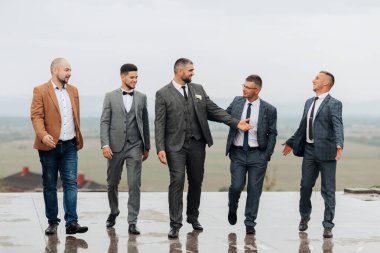 Takım elbiseli ve kravatlı beş kişilik bir grup ıslak bir yüzey üzerinde duruyor, muhtemelen bir çatı. Hepsi gülümsüyor ve birbirlerinin arkadaşlığından zevk alıyor gibi görünüyorlar. Sahne neşeli ve dostane.