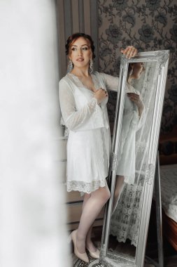 Beyaz elbiseli bir kadın aynanın önünde duruyor. Bir çift küpe ve kolye takıyor.