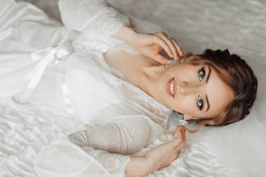 Beyaz bornozlu bir kadın yatakta yatıyor. Küpe takıyor ve yüzünde bir gülümseme var.