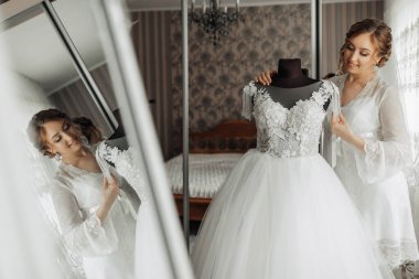 Bir kadın aynanın önünde elinde gelinlik tutuyor. Elbise beyaz ve çiçek desenli. Kadın beyaz bir elbise giyiyor ve aynada kendine bakıyor.