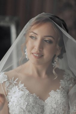 Bir gelin peçe takıyor ve gülümsüyor. Duvak beyaz ve dantel bir tasarımı var. Gelinin elbisesi de beyaz ve dantelli.
