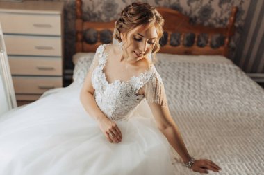 Gelinlik giymiş bir kadın yatakta oturuyor. Gülümsüyor ve kameraya bakıyor.