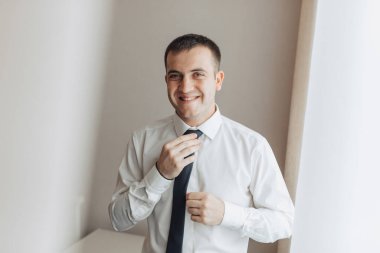 Beyaz gömlek ve siyah kravat takan bir adam var. Gülümsüyor ve kameraya bakıyor.