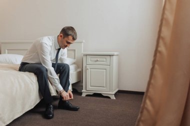 Bir adam yatakta oturuyor ve ayakkabılarını tamir ediyor. Adamın takım elbise ve kravat giydiği ama resmi bir ortamda olmadığı için resmin rahat ve rahat bir havası var.