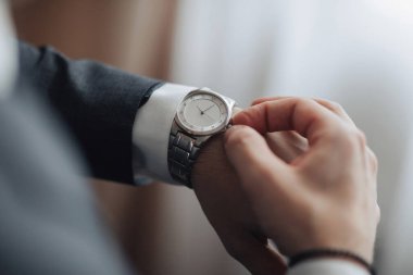 Bir adam saat takıyor ve ona bakıyor. Saat gümüşten ve siyah bir yüzü var.