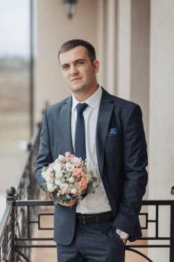 Elinde bir buket çiçek tutan takım elbiseli bir adam. Gülümsüyor ve kameraya bakıyor.
