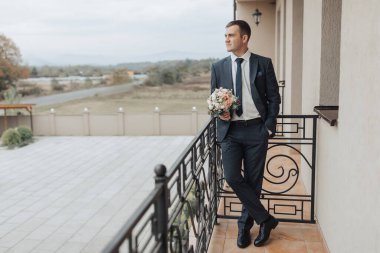 Takım elbiseli ve kravatlı bir adam elinde bir buket çiçekle balkonda duruyor. Manzara sakin ve huzurlu, adamın güzel manzaraya bakışıyla.