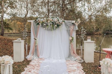 Bir parkta düğün töreni düzenleniyor. Gelin ve damat çiçeklerle süslenmiş beyaz bir kemerin önünde duruyorlar. Kemer beyaz bir perdeyle çevrili.