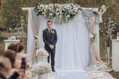 Üzerinde çiçekler olan beyaz bir perdenin önünde duran bir adam. Takım elbise ve kravat takıyor.