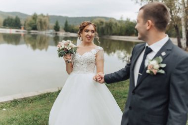 Gelin ve damat gölün kenarında, gelin elinde bir buket çiçek tutuyor. Adam kravat takıyor ve kadın beyaz bir elbise giyiyor. Sahne romantik ve resimli.