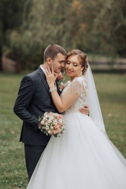 Bir gelin ve damat parkta fotoğraf için poz veriyorlar. Gelin beyaz bir elbise giyiyor ve damat bir takım elbise giyiyor. El ele tutuşup gülümsüyorlar, mutlu ve aşık görünüyorlar.