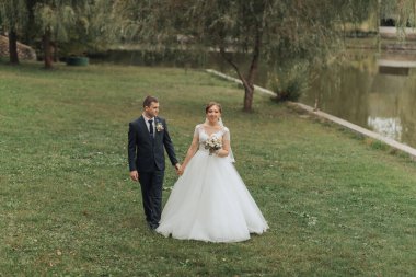 Bir gelin ve damat parkta birlikte yürüyorlar. Gelin beyaz bir elbise giyiyor ve damat bir takım elbise giyiyor. El ele tutuşuyorlar ve mutlu görünüyorlar.