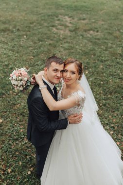 Bir gelin ve damat tarlada fotoğraf çektirmek için poz veriyorlar. Gelin beyaz bir elbise giyiyor ve damat bir takım elbise giyiyor. İkisi de gülümsüyor ve birbirlerine sarılıyorlar. Sahne mutlu ve romantik.