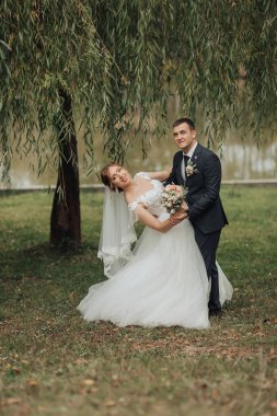 Bir gelin ve damat parkta fotoğraf için poz veriyorlar. Gelin beyaz bir elbise giyiyor ve damat bir takım elbise giyiyor. Elinde bir buket ve bir buket tutuyorlar.