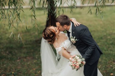 Parkta bir gelin ve damat öpüşüyor. Gelin beyaz bir elbise giyiyor ve damat da takım elbise giyiyor.