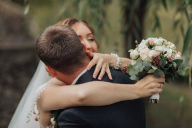 Bir erkek ve bir kadın birbirlerine sarılıyorlar, adam elinde bir buket çiçek tutuyor.